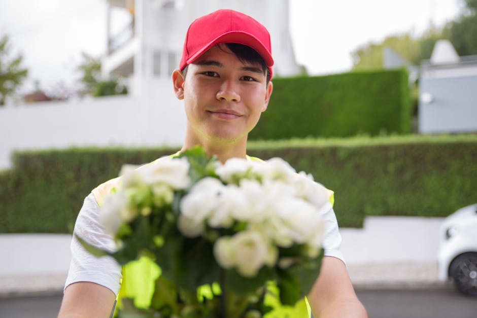 A delivery person wearing a red cap and a reflective yellow vest is smiling and holding out a bouquet of fresh white roses and green foliage towards the camera. The bouquet is wrapped in clear celloph