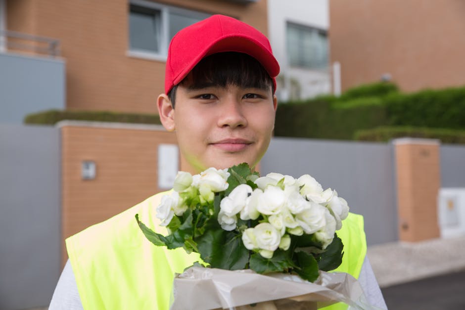 A young delivery person wearing a red cap and high-visibility yellow vest holds a bouquet of white roses with lush green leaves, wrapped in white floral paper. The person stands outdoors in front of m
