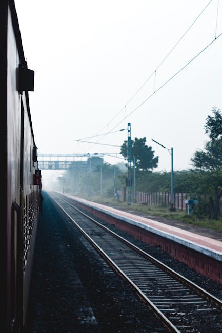 This image depicts the exterior of a train carriage, partially visible on the left side, with the window framing the scene outside. The train appears to be stationary at a platform, with the view exte