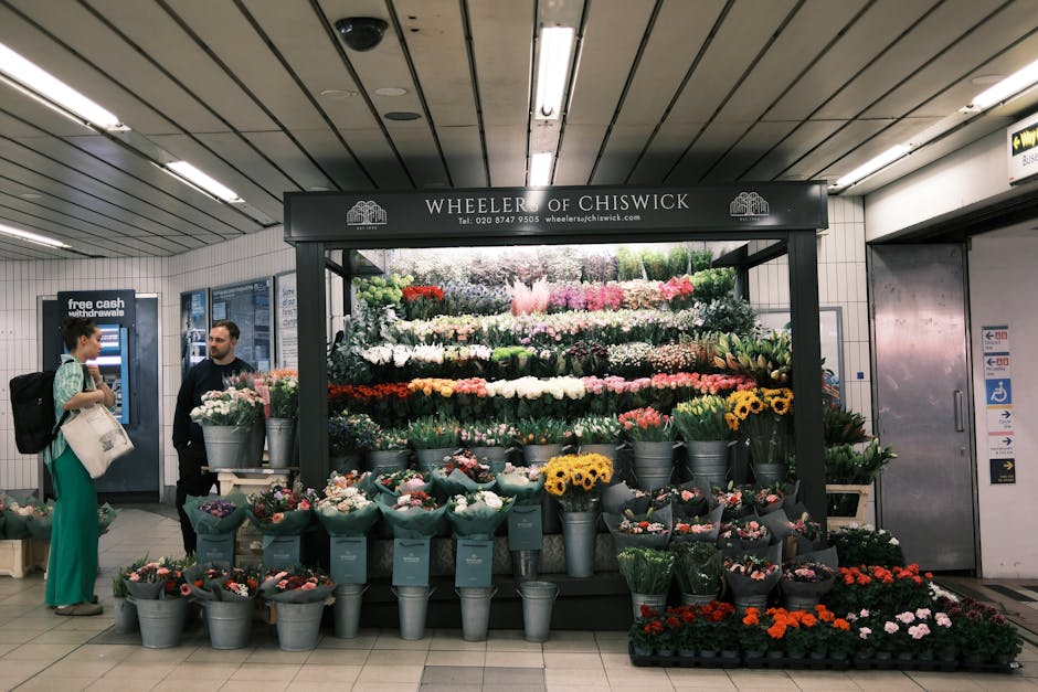 A vibrant flower display at Wheeler of Chiswick, set up in an indoor space beneath a ceiling with linear metal panels, featuring a variety of fresh flowers arranged in tiered layers. The arrangement i