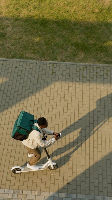 An aerial view of a person riding a white electric scooter on a paved pathway with rectangular beige bricks. The individual is wearing a white jacket, beige pants, and a white face mask, carrying a la